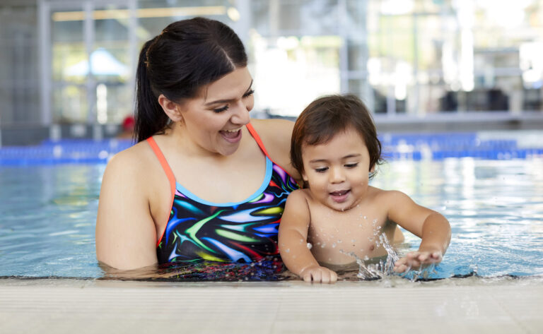 Photo of mother smiling whilst holding her baby along the pool edge as the baby lightly splashes their hands in the water.